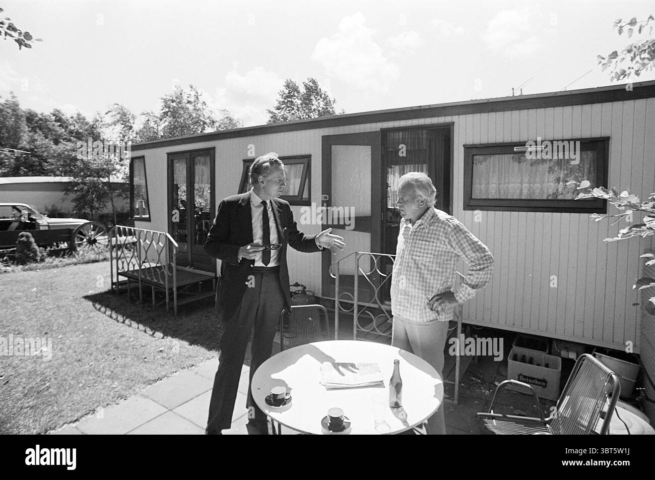 Duinrell camping in Wassenaar Kampen campsites camp site camping camping hut Tourism tourists, Whizgle News, Dutch Desk, The Netherlands, 1950 - 2000 on 26-07-1974. The image shows these topics. In a sunlit outdoor setting, two men engage in conversation in front of a modest mobile home. One man, dressed in a dark suit with a tie, stands animatedly, gesturing with his hands as he speaks. His hair is slicked back, emphasizing a focus on communication. The second man, wearing a light checkered shirt, leans slightly against the table, appearing more relaxed yet engaged in the dialogue. The mobile Stock Photo