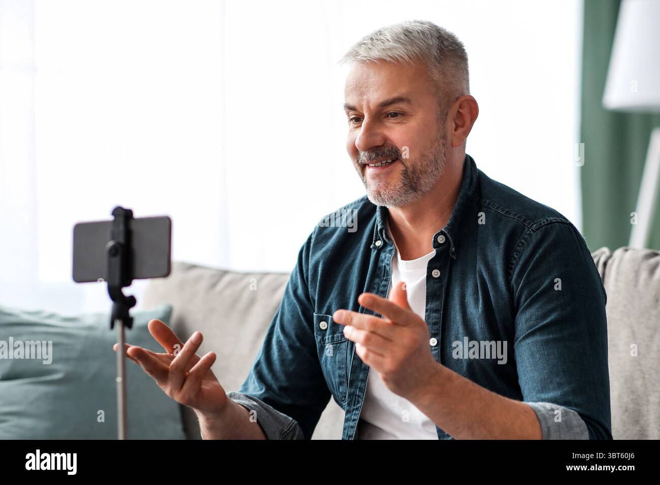 Attractive mature man broadcasting from home, using smartphone on tripod Stock Photo