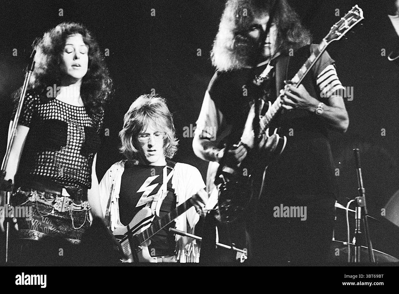 Holland Pop Festival in the Kralingse bos which has gone down in history as the Dutch version of the American Woodstock festival. With performances by The Byrds Mungo Jerry Canned Heat and Jefferson Airplane Music Rotterdam, Whizgle News, Dutch Desk, The Netherlands, 1950 - 2000 on 26-06-1970. These are the topics in the image. The scene captures a dynamic moment in a musical performance, featuring three individuals drenched in the energy of the stage. To the left, a woman stands confidently, her long dark hair cascading down her shoulders. She wears a revealing black mesh top paired with fitt Stock Photo