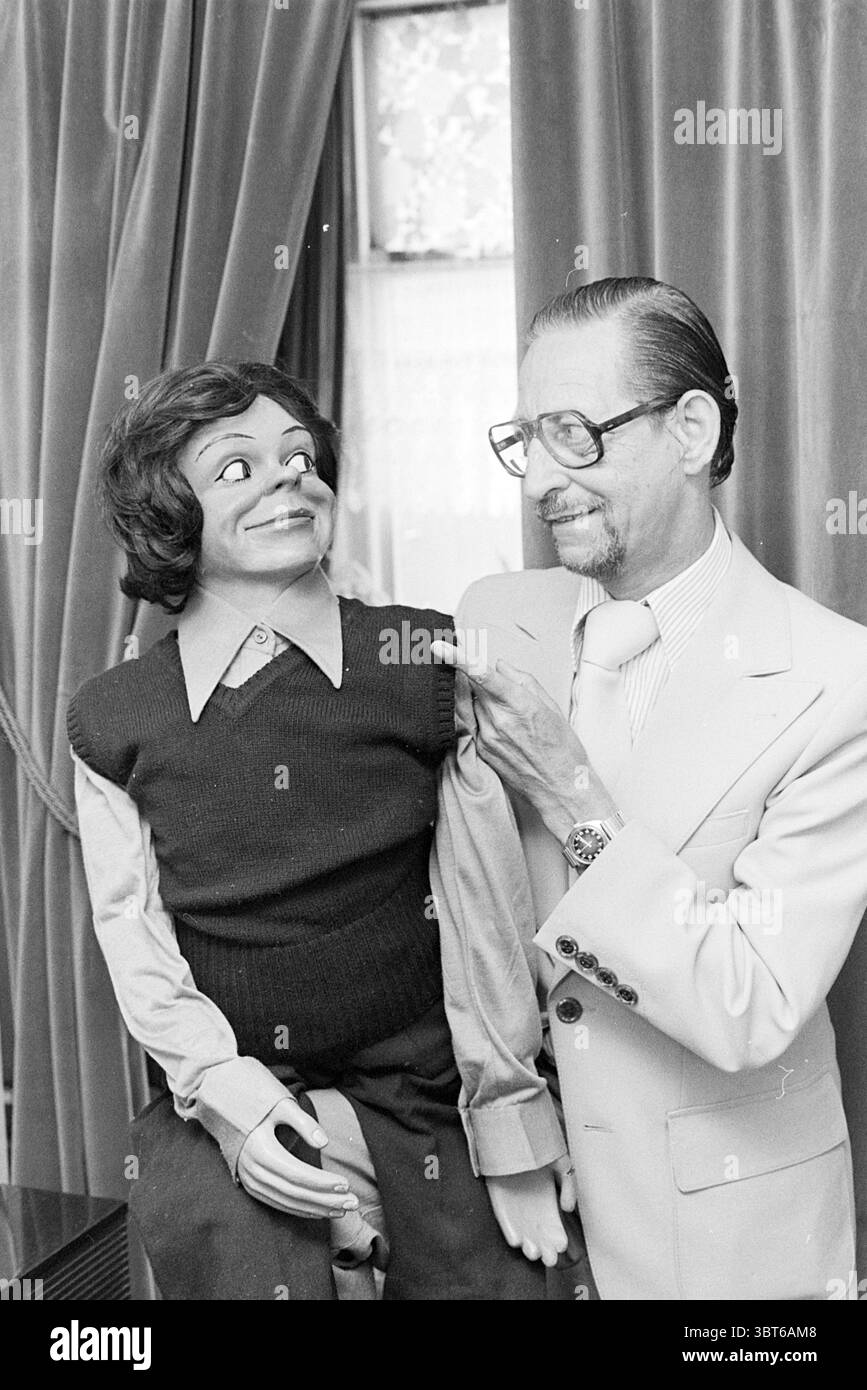 Daan Vrolijk. Persons, Whizgle News, Dutch Desk, The Netherlands, 1950 - 2000 on 07-07-1977. These topics appear in the image. In a warmly lit interior, a man stands next to a ventriloquist's dummy, creating an engaging and whimsical scene. The man, dressed in a light-colored suit with a tie, sports round glasses and has neatly combed hair. His expression conveys a mix of pride and amusement as he gestures towards the dummy. The dummy, with a painted face characterized by exaggerated features, has wavy brown hair, a broad smile, and lively eyes, suggesting an animated personality. It is dresse Stock Photo