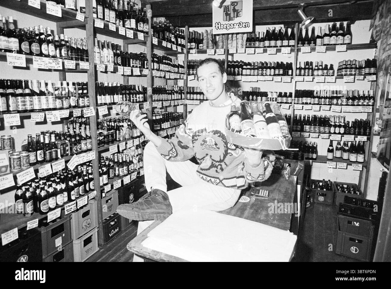 Beer shop Deux Bières, Whizgle News, Dutch Desk, The Netherlands, 1950 - 2000 on 27-01-1993. These are the elements in the image. The scene features a bustling beverage shop, filled with numerous bottles of various sizes lining the shelves behind a central figure. The shelves create an organized yet slightly chaotic backdrop, showcasing an impressive array of labels and colors, predominantly dark hues from the bottles amid bright accents from the labels. In the foreground, a person sits confidently on a well-worn wooden counter, contributing a human element to the otherwise densely packed spac Stock Photo