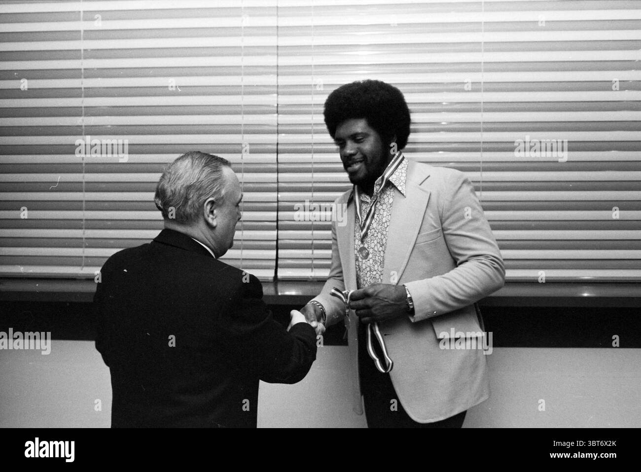 Baseball and softball awards ceremony Baseball Awards Ceremony, Whizgle News, Dutch Desk, The Netherlands, 1950 - 2000 on 14-11-1980. The image includes these topics. The scene captures a moment with two men engaging in a handshake, conveying a sense of mutual respect and achievement. One man, dressed in a formal dark suit, appears older, his hair graying, and showcases a serious expression as he extends his hand. The other man, sporting an expressive, voluminous afro, is clad in a stylish light-colored blazer over a patterned shirt, suggesting a vibrant personality. He wears a medal around hi Stock Photo