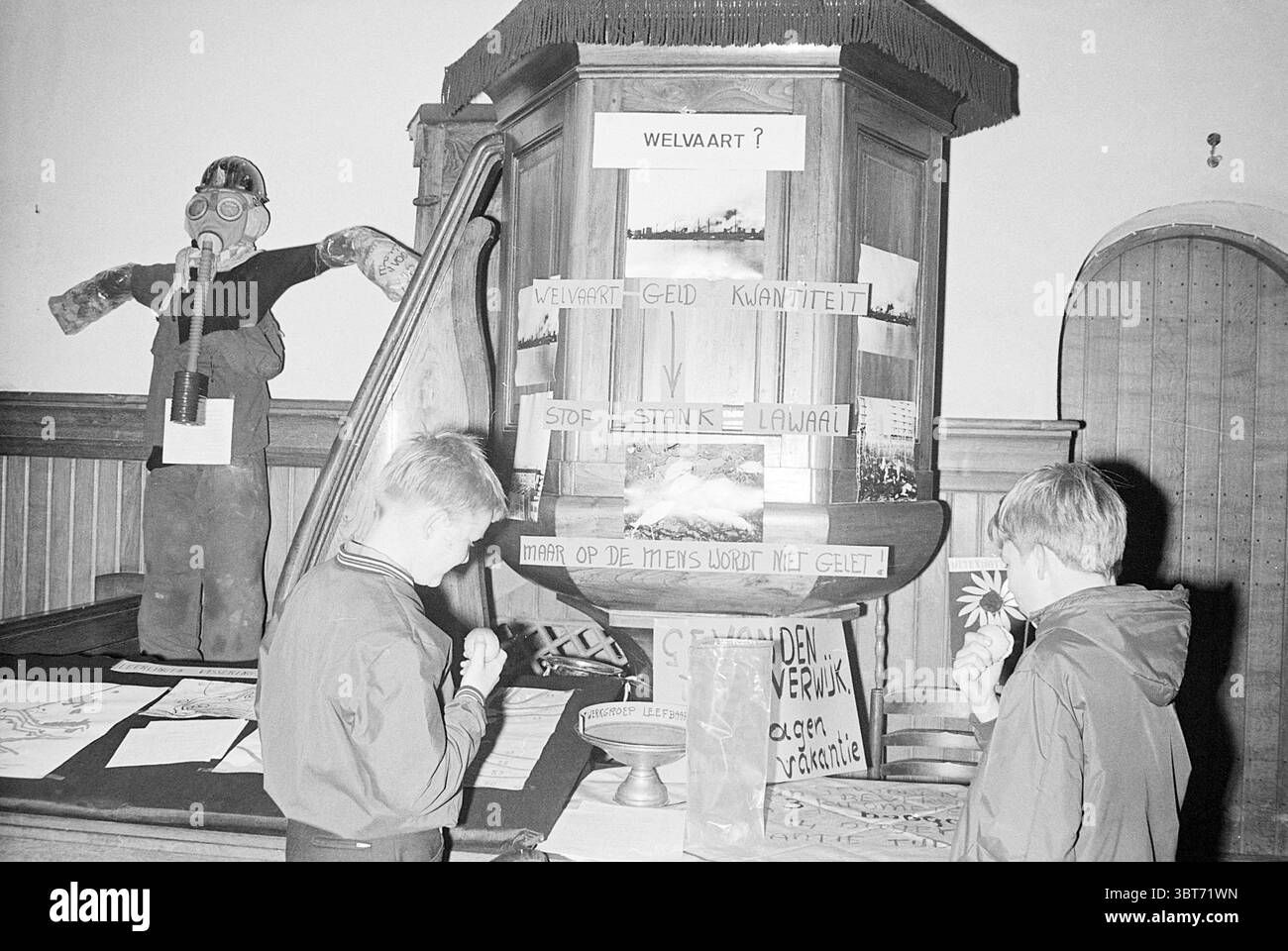 Opening Livability Exhibition Opening openings commissioning, Whizgle News, Dutch Desk, The Netherlands, 1950 - 2000 on 19-10-1970. The image contains these topics. The scene takes place in a room with a historical or educational atmosphere, characterized by wood-paneled walls that convey a sense of warmth. In the center, a wooden display stand holds various items, drawing the attention of two children standing close by. The display features a prominent, elaborately carved structure, likely a pulpit or lectern, topped with a decorative fringe. It is adorned with papers and objects, hinting at Stock Photo
