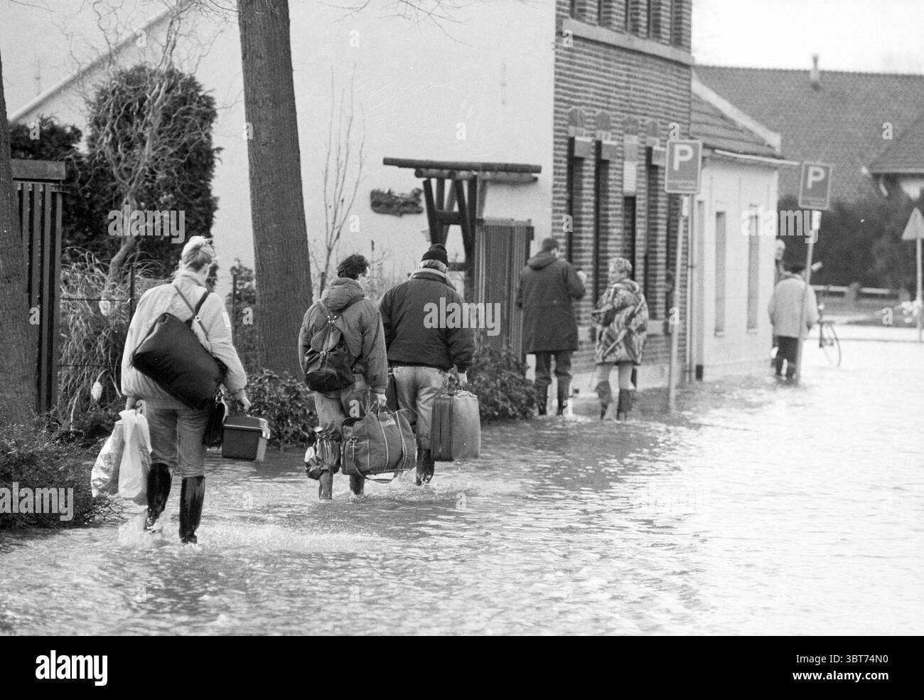 Flood Limburg Roermond Roermond, Whizgle News, Dutch Desk, The Netherlands, 1950 - 2000 on 22-12-1993. These topics are shown in the image. The scene captures a somber moment in an urban setting affected by flooding. In the foreground, several individuals are wading through murky water that reaches up to their mid-calf, indicating significant inundation. They are dressed in a mix of casual and rugged clothing suitable for the weather, with some wearing waterproof boots, while others carry bags and personal items, suggesting a hurried evacuation. The overall color palette is muted, dominated by Stock Photo