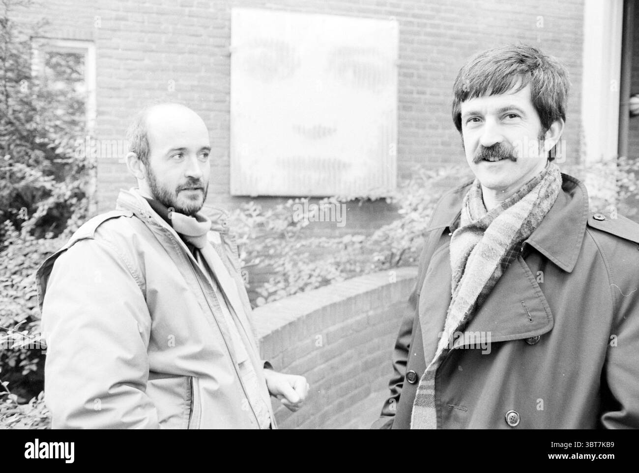 Joop Glasbergen and Mr. Mielgas Foundation Bavo Foundation, Whizgle News, Dutch Desk, The Netherlands, 1950 - 2000 on 03-11-1986. The image shows these topics. The scene features two men standing outdoors against a backdrop of a textured brick wall, which has an abstract representation of a face, subtly integrated into the background. The first man on the left has a shaved head and a short beard, exuding a thoughtful demeanor. He is dressed in a light-colored jacket and has a scarf around his neck, giving him a cozy and somewhat informal appearance. The second man, positioned on the right, has Stock Photo