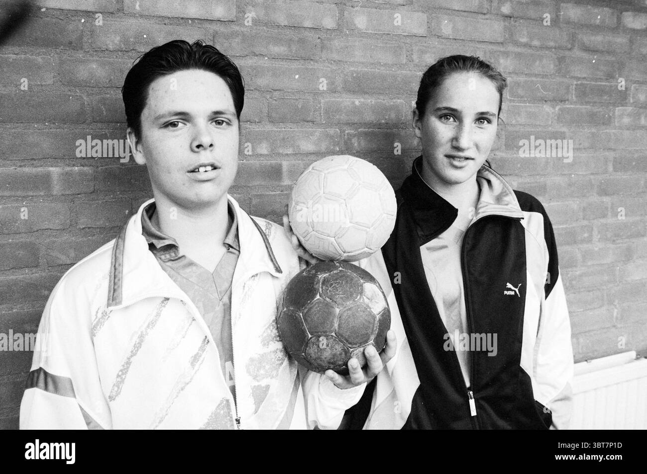 Suz. Jerphanion and Rene Greve Handball, Whizgle News, Dutch Desk, The Netherlands, 1950 - 2000 on 13-11-1993. The image contains these topics. In this scene, two young individuals stand closely together against a backdrop of textured brick, creating a stark and engaging environment. The first person on the left has dark hair, styled neatly, wearing a light-colored sporty jacket that features distinct lines and patterns, giving off a retro vibe. Their expression is somewhat serious, conveying a sense of determination or focus. The second individual, on the right, has longer hair pulled back an Stock Photo