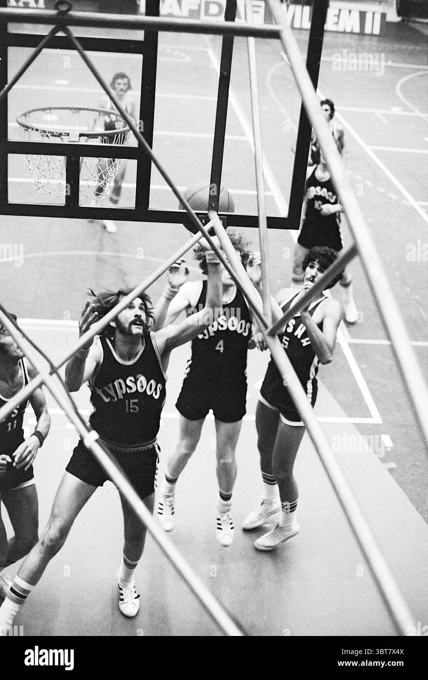 Basketball match: Typsoos - Bosma., Whizgle News, Dutch Desk, The Netherlands, 1950 - 2000. The image includes these topics. In a dynamic indoor sports setting, a group of athletes is engaged in an intense basketball game. The scene is filled with movement and energy. Several players, adorned in matching dark jerseys with the number '15' prominently displayed, are in the midst of a competitive play under the basketball hoop. The player wearing jersey number '15' is leaping upward, arms extended, as she attempts to shoot the basketball towards the hoop. Her hair is in motion, accentuating the e Stock Photo