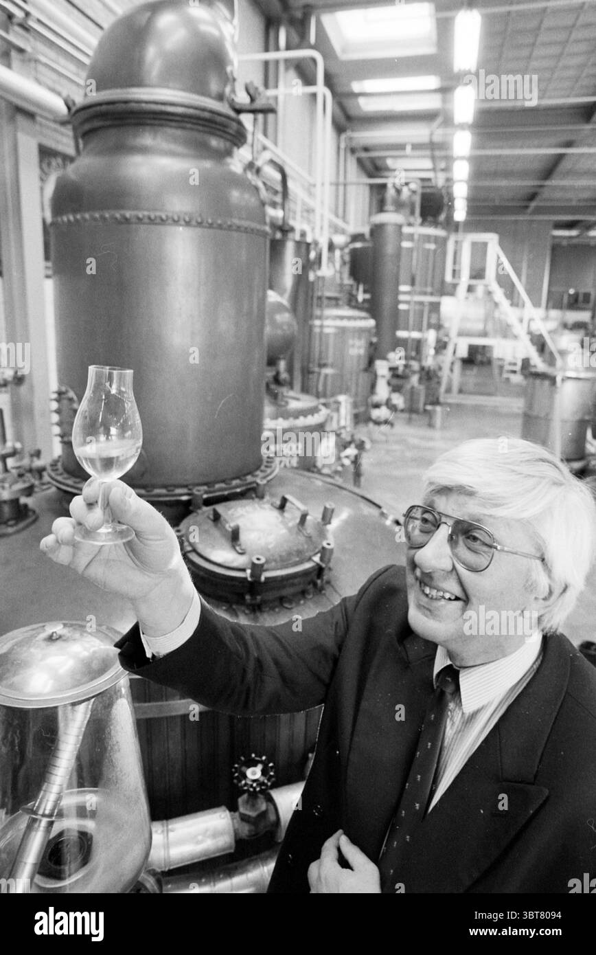 Master Distiller Bols Nieuw-Vennep Lucas Bolsstraat, Whizgle News, Dutch Desk, The Netherlands, 1950 - 2000 on 16-03-1992. These are the elements in the image. In a bustling industrial space, a gentleman stands prominently in the foreground, exuding a sense of pride and accomplishment. He has a light complexion, and his neatly styled hair is a silver-gray hue, complementing his sharp suit, which is a classic dark color. The suit is accessorized with a tie that suggests a subtle pattern, adding a touch of personality to his formal appearance. In his right hand, he holds a small, elegant glass d Stock Photo