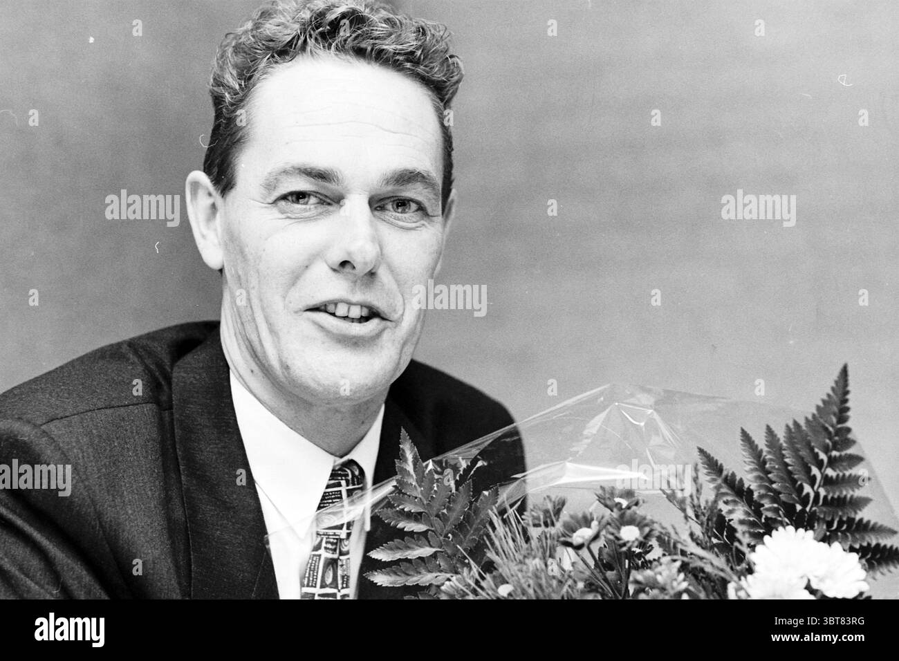Portrait man with bunch of flowers, Whizgle News, Dutch Desk, The Netherlands, 1950 - 2000 on 12-1997. These topics appear in the image. The scene features a man seated at a table, exuding a warm and approachable demeanor. He has short, neatly styled hair, showcasing a natural wave, and his expression is friendly, with a slight smile that highlights his engaging personality. His attire consists of a well-fitted dark suit, complemented by a tie adorned with a geometric pattern in contrasting colors, adding a touch of sophistication to his look. In his hands, he holds a bouquet of flowers, which Stock Photo