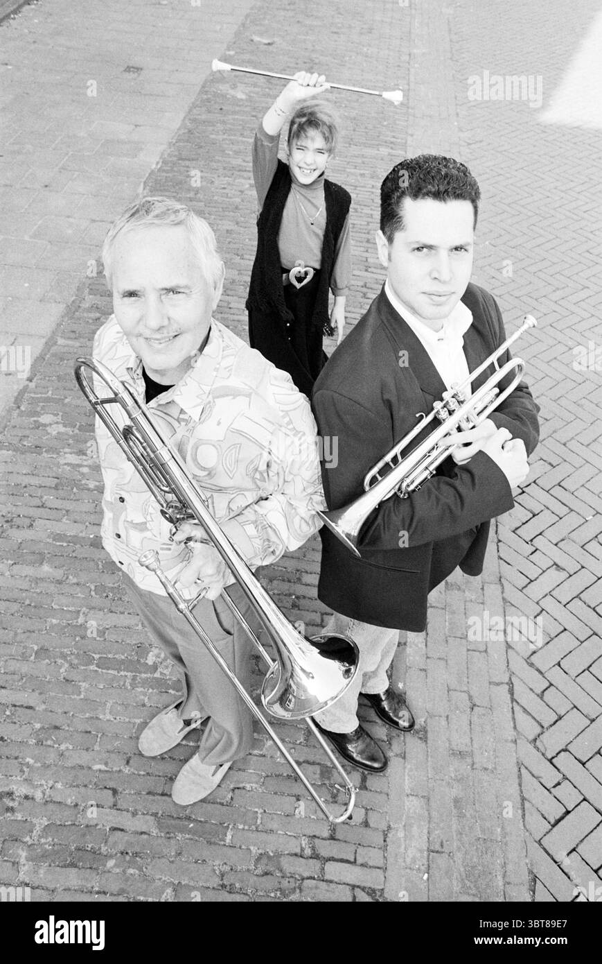 Jr. + Sr. Calis family, Whizgle News, Dutch Desk, The Netherlands, 1950 - 2000 on 26-03-1994. These are the topics in the image. In a visually striking scene, three musicians are positioned in a dynamic arrangement, creating a sense of movement and energy. The primary subjects, a male trombonist and a male trumpeter, are standing closely together, both holding their instruments confidently. The trombonist, dressed in a patterned shirt that boasts swirling designs, has gray hair and an engaging expression that suggests a playful personality. His stance is relaxed yet poised, with the trombone h Stock Photo