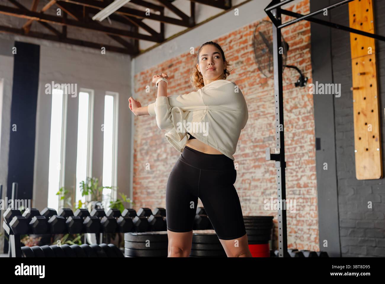 Woman stretching shoulder across chest in fitness studio with fitness watch dumbbells pullup rig Stock Photo