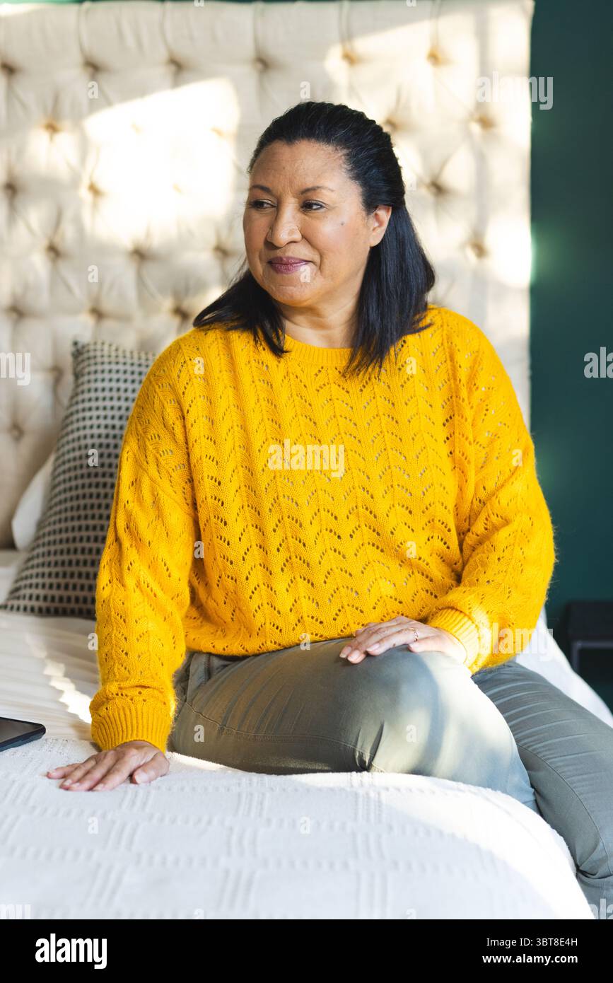 Woman sitting on bed with decorative pillows at home wearing mustard yellow sweater with smartphone Stock Photo