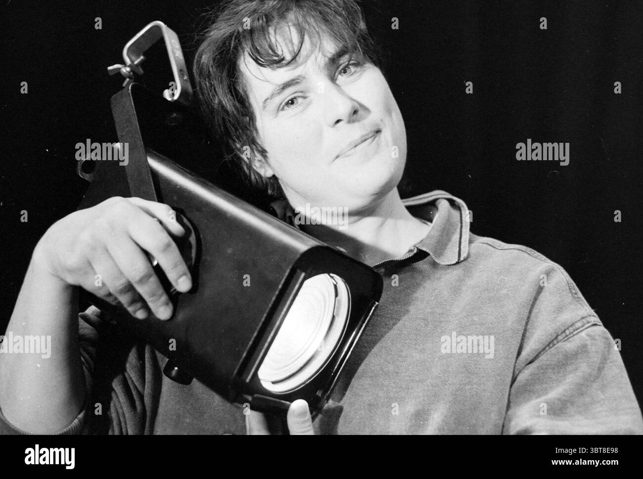 Isabel Hamer lighting technician, Whizgle News, Dutch Desk, The Netherlands, 1950 - 2000 on 02-05-1991. The image shows these topics. The scene features a woman with a confident and playful expression, highlighting a warm personality. She has short, tousled hair and is positioned slightly off-center, drawing attention to her engaging smile. The woman is holding a spotlight, a large, black object with a shiny circular lens, which adds a sense of depth and interest to the composition. The background is dark, likely a deep black, creating a stark contrast that emphasizes her figure and the spotli Stock Photo