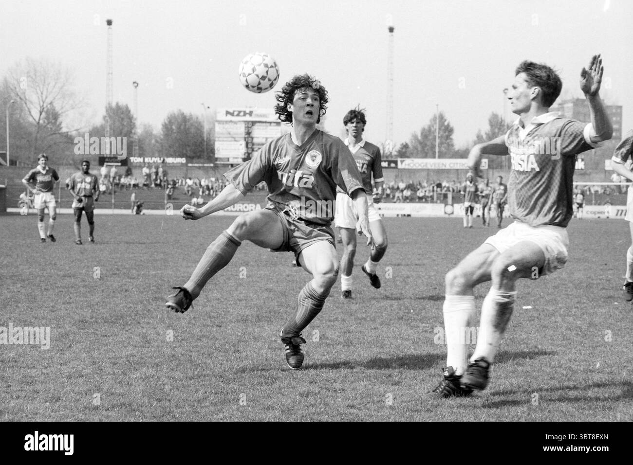 Haarlem - AZ, Whizgle News, Dutch Desk, The Netherlands, 1950 - 2000 on 13-04-1991. These are the topics in the image. The scene captures an intense moment on a grassy soccer field under a clear sky. Two players dominate the foreground; one, wearing a dark jersey, is airborne, having just executed a graceful maneuver to control the ball with his feet. His hair is curly and windswept, emphasizing the dynamic motion of the play. The other player, clad in a lighter jersey, is in a defensive stance, his body slightly bent and arms outstretched as he prepares to intercept or react to the airborne p Stock Photo