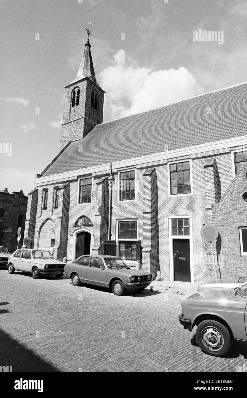 Restored Walloon Church Benijnenhof Restoration Haarlem Begijnhof The Netherlands, Whizgle News, Dutch Desk, The Netherlands, 1950 - 2000 on 04-05-1982. The image includes these topics. The scene depicts a quaint street lined with vintage cars parked alongside a charming brick building with an architectural style that hints at historical significance. The building features a tall, narrow steeple rising above the roofline, casting a slender shadow on the cobblestone pavement below. Its red-brick facade is interspersed with large, rectangular windows framed in white, which allow glimpses of the Stock Photo