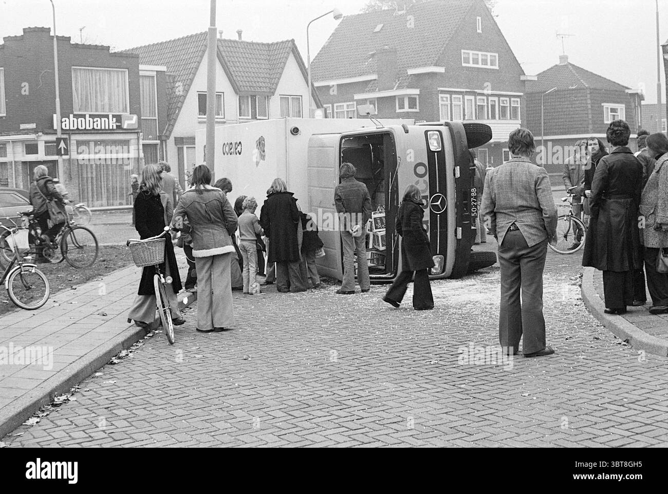 Accident overturned truck, Whizgle News, Dutch Desk, The Netherlands, 1950 - 2000 on 10-1976. These topics are shown in the image. The scene depicts a bustling urban street corner where a white delivery truck has overturned onto its side. The truck's large wheels are facing upward, and its door is ajar, suggesting a hurried escape or unexpected event. Surrounding the truck is a crowd of onlookers, a mix of adults and children, who are gathered to witness the unfolding situation. Many individuals appear curious and are peering inside the truck or discussing the incident amongst themselves. The Stock Photo