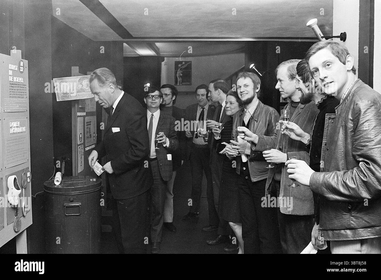 Opening of the exhibition Theater Opening openings opening, Whizgle News, Dutch Desk, The Netherlands, 1950 - 2000 on 06-03-1969. These are the elements in the image. The scene unfolds in a dimly lit interior, creating a somewhat intimate and nostalgic atmosphere. To the left, a well-dressed man in a dark suit leans slightly forward, intently focused on a tall, cylindrical object that resembles a trash can or a similar receptacle. He appears to be engaging with it, perhaps in a playful or curious manner. A group of individuals stands to the right, casually positioned as they observe the man. T Stock Photo