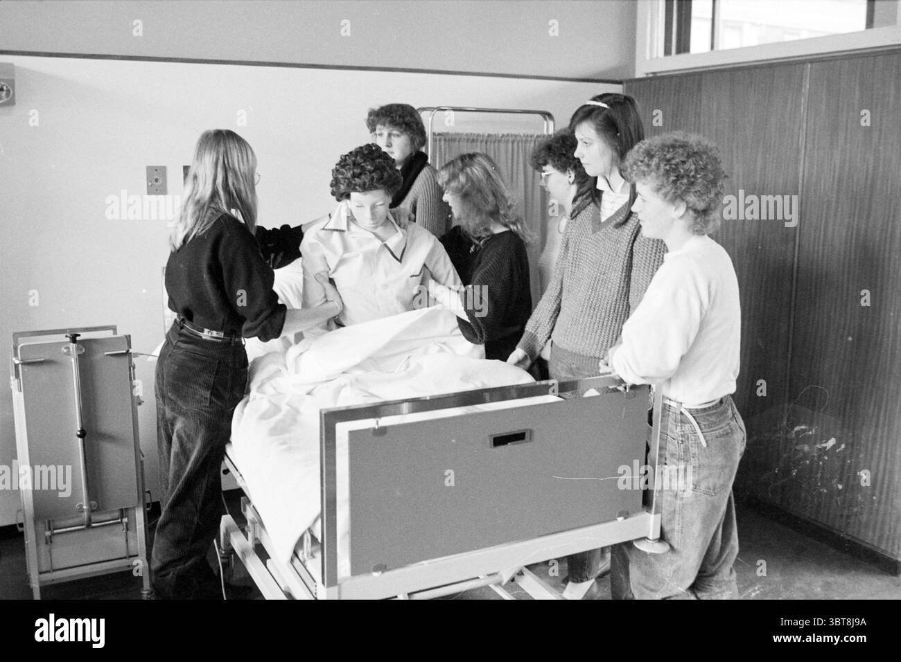 Training center IJmuiden Zeeweg hospital IJmuiden The Netherlands, Whizgle News, Dutch Desk, The Netherlands, 1950 - 2000 on 02-04-1984. The image includes these topics. In a well-lit room, a group of six individuals is gathered around a hospital bed. The setting appears to be a medical training environment, characterized by neutral-colored walls and a large window allowing natural light to infuse the space. The individuals display a variety of hairstyles and clothing styles, indicating a casual yet focused atmosphere. One person, positioned at the foot of the bed, is wearing a white coat, whi Stock Photo