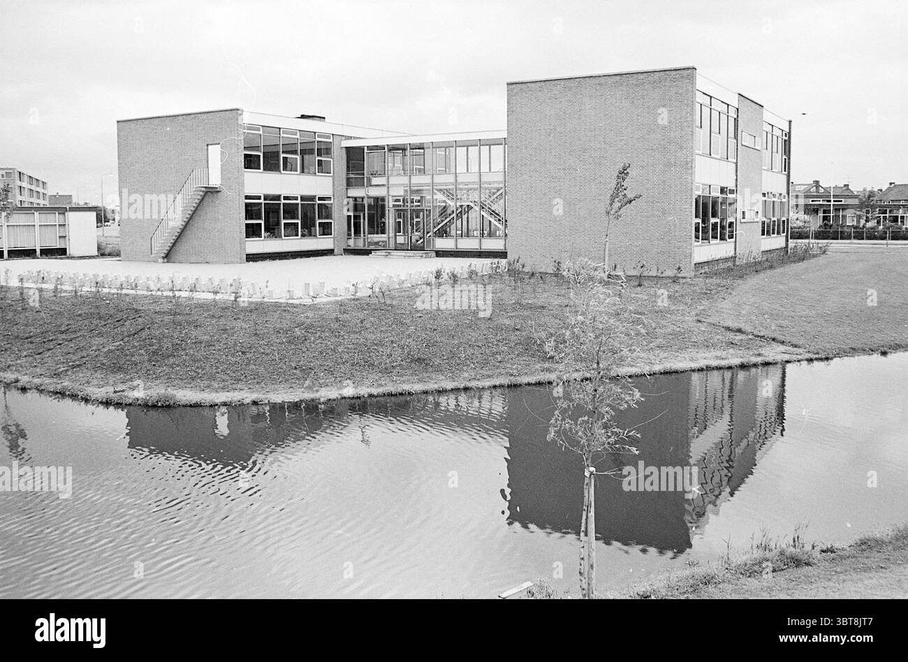 School in Zwanenburg Houses and house construction, Whizgle News, Dutch Desk, The Netherlands, 1950 - 2000 on 27-07-1966. These are the topics in the image. The scene features a modern building characterized by its geometric shapes and expansive glass windows, allowing for a clear view of its interior. The structure has a combination of brick and glass, with a slightly industrial feel due to its straightforward lines and functionality. To the side, an external staircase leads up to a second floor, emphasizing the building's multi-level design. Surrounding the structure is a well-maintained gra Stock Photo