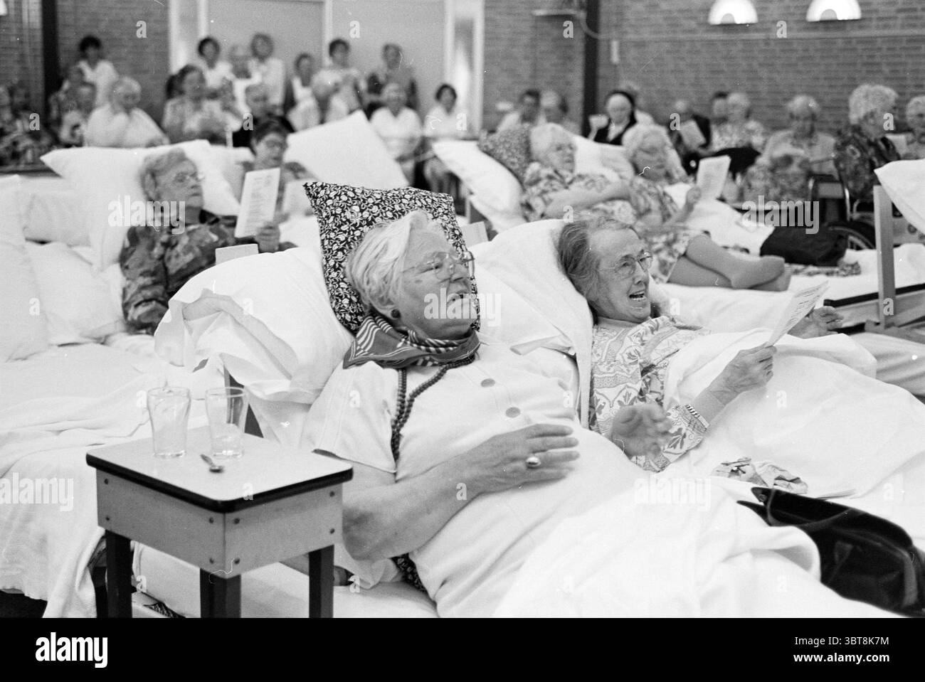 Recreation room with people in need plus nursing staff (performance by children's choir), Whizgle News, Dutch Desk, The Netherlands, 1950 - 2000 on 06-1996. These topics appear in the image. The scene captures a large room filled with elderly women, each settled in beds that are closely arranged, showcasing an atmosphere of community. The setting has soft, neutral-toned walls, accentuated by the warm glow of overhead lighting that adds a cozy, intimate feeling to the environment. In the foreground, a couple of women are prominently featured lounging on their beds, wearing patterned headscarves Stock Photo