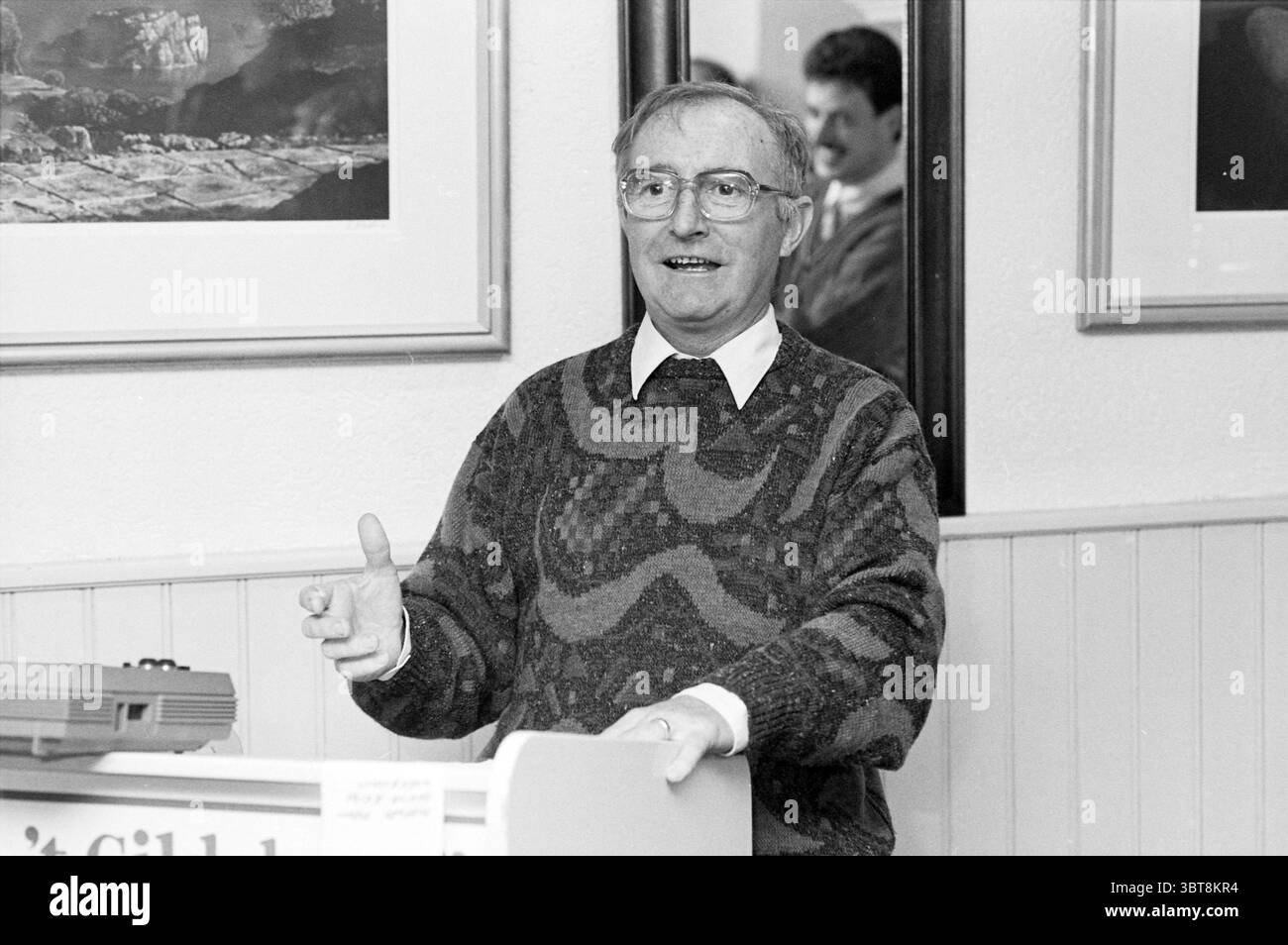 Reception Mr. Harnborn guildhall Beverwijk N.A.M. Beverwijk The Netherlands, Whizgle News, Dutch Desk, The Netherlands, 1950 - 2000 on 28-10-1987. These are the elements in the image. The scene depicts a man actively engaging with an audience, standing at a lectern adorned with a subtle design. He has a friendly expression, his smile suggesting warmth and approachability, as he gestures with one hand to emphasize his point. His attire consists of a patterned sweater in muted tones, which offers a cozy, casual feel to the setting. Behind him, several framed artworks hang on the wall, showcasing Stock Photo