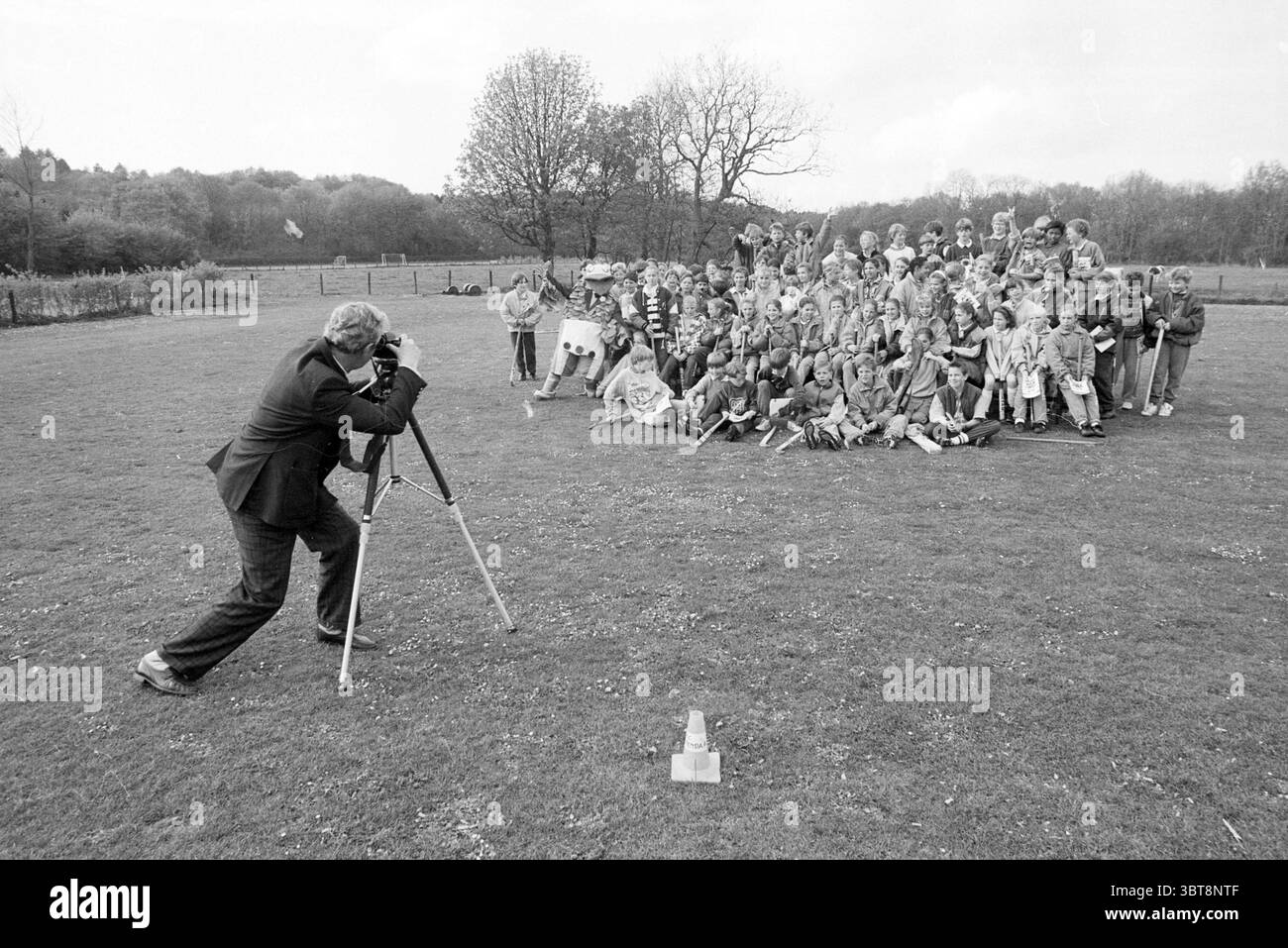 Presentation of diplomas in hockey B'daal Hockey Presentation Bloemendaal, Whizgle News, Dutch Desk, The Netherlands, 1950 - 2000 on 27-04-1990. The image shows these topics. The scene depicts a large group of children gathered together outdoors, smiling and posing for a photograph. They are arranged in several rows, with the front row crouching and the back row standing. The children are dressed in light jackets and pants, showcasing a variety of colors, including reds, blues, yellows, and greens. In the foreground, a man, possibly the photographer, is focused on capturing the moment using a Stock Photo