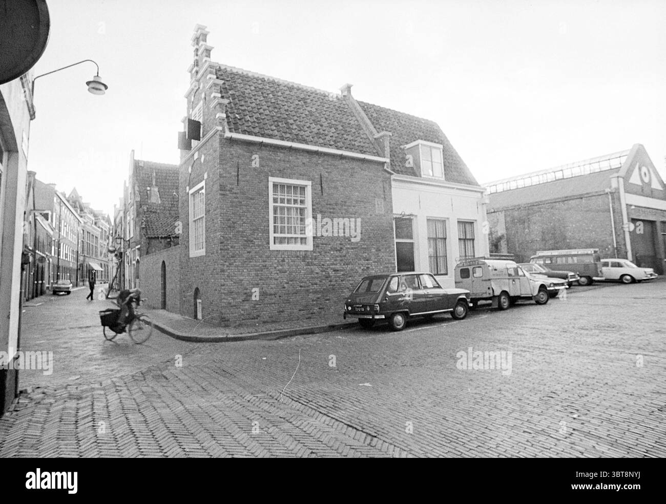 Begijnhof building Exterior Haarlem Begijnhof The Netherlands, Whizgle News, Dutch Desk, The Netherlands, 1950 - 2000 on 15-09-1972. These are the elements in the image. The scene features a quiet, cobblestone street corner in a quaint town, accentuated by historical architectural elements. To the left, a narrow passage leads into a cluster of old brick houses, their facades showcasing a sturdy, rustic charm. The two-story buildings, with their steeply pitched roofs, feature white window frames and small, diamond-paned glass windows. In the foreground, a cyclist passes by, slightly blurred to Stock Photo