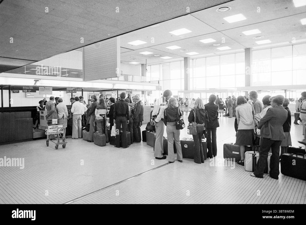Holiday crowds Schiphol Busy Holidays holidaymakers, Whizgle News, Dutch Desk, The Netherlands, 1950 - 2000 on 01-07-1977. The image contains these topics. In a bustling airport terminal, a long line of travelers stretches toward a check-in counter. The scene is characterized by a monochromatic color palette, dominated by shades of gray and black, contributing to a nostalgic atmosphere. People of various ages and backgrounds stand in the queue, many holding black and dark-colored suitcases, some adorned with stickers from different destinations. A few individuals are dressed in smart-casual at Stock Photo