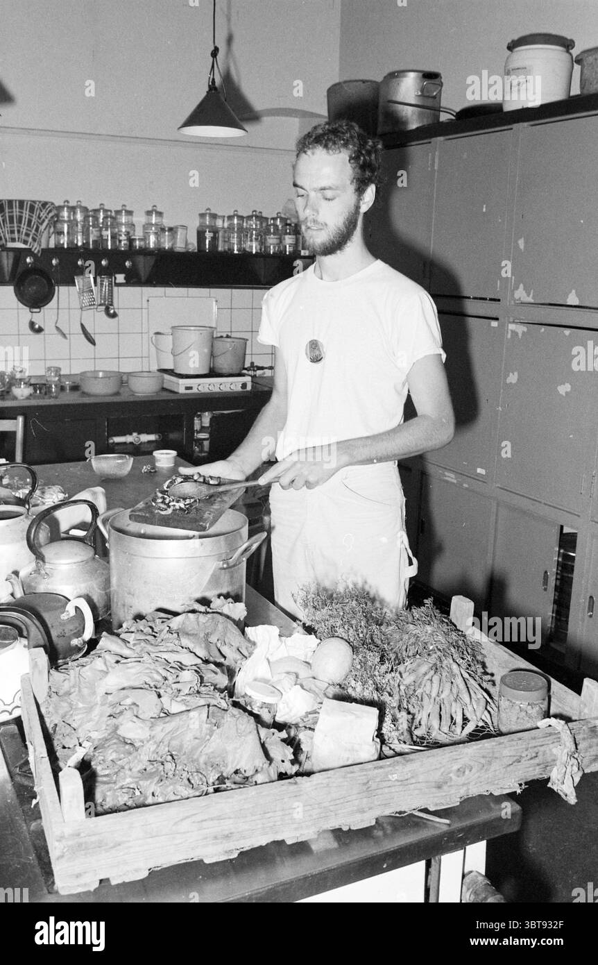 Kitchen (vegetarian) Academy of Natural Medicine Bloemendaal Vegetarian Bloemendaal, Whizgle News, Dutch Desk, The Netherlands, 1950 - 2000 on 28-06-1982. These are the topics in the image. The scene reveals a bustling kitchen environment filled with various elements that contribute to its lively atmosphere. In the foreground, a wooden table is laden with a variety of fresh vegetables and ingredients: leafy greens, a bulbous onion, and herbs, their textures and colors contrasting against one another. The arrangement is somewhat casual, suggesting a hurried yet vibrant preparation process. To t Stock Photo