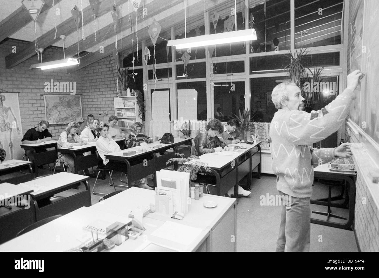 That. Meyermans teaches parents at Moerberg School Schools, Whizgle News, Dutch Desk, The Netherlands, 1950 - 2000 on 10-02-1987. These are the elements in the image. The scene depicts a classroom setting, characterized by a mix of natural and artificial lighting illuminating the space. Large windows line one side of the room, allowing soft daylight to filter in, casting gentle shadows on the floor and enhancing the overall atmosphere. In the foreground, a teacher stands confidently at the front, engaging with the class. He has light-colored hair and is wearing a sweater with a distinct zigzag Stock Photo