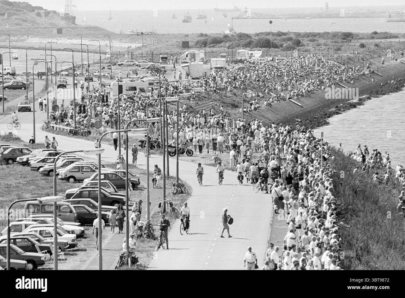Departure sail ships IJmuiden The Netherlands, Whizgle News, Dutch Desk, The Netherlands, 1950 - 2000 on 14-08-1990. These topics appear in the image. The scene captures a vibrant outdoor event, bustling with numerous people gathered along a scenic waterfront. The setting features a long, winding path bordered on one side by a lush green area and the sparkling water on the other. A diverse crowd, dressed in light, casual summer attire, stretches along the path. Some individuals stand together in conversation, while others stroll leisurely or ride bicycles, creating a dynamic atmosphere full of Stock Photo