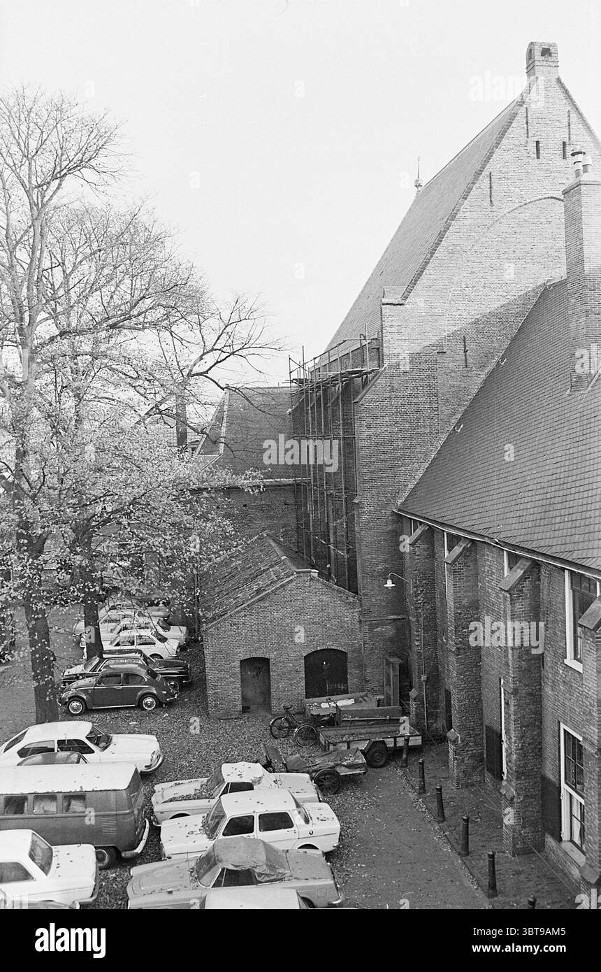 Exterior Begijnhof 20 Exterior Haarlem Begijnhof The Netherlands, Whizgle News, Dutch Desk, The Netherlands, 1950 - 2000 on 16-10-1970. The image includes these topics. The scene depicts a historical building with a rustic charm, characterized by its textured brick facade and distinct architectural angles. The structure has a steeply pitched roof, complemented by nearby scaffolding indicating ongoing repairs or renovations. In the foreground, a cluster of vehicles is parked closely together, showcasing a variety of older model cars rendered in muted tones of white, gray, and black. The vehicle Stock Photo