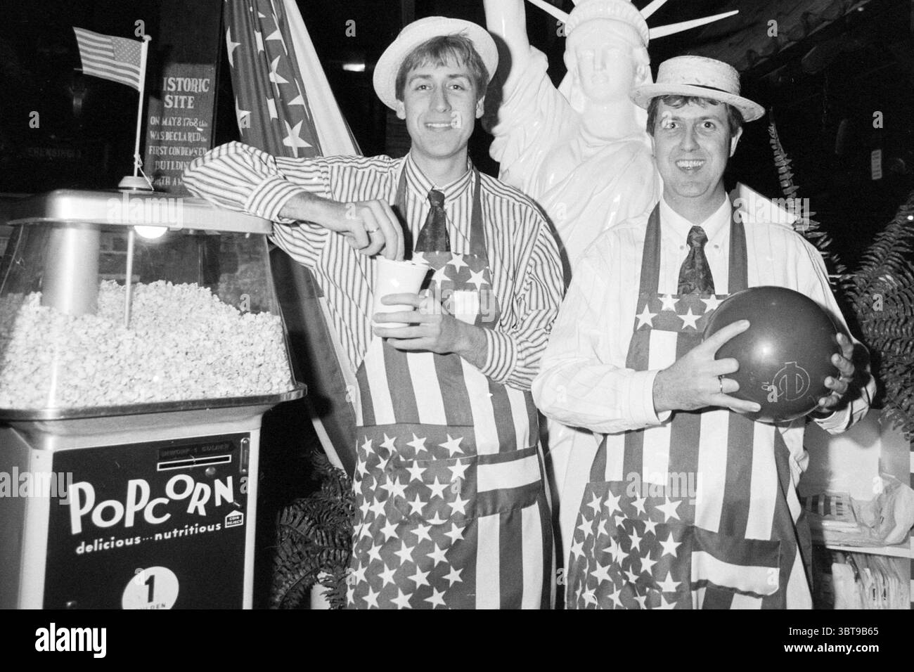 Gebr. Claus due to USA week Claus Party House Bowling Haarlem The Netherlands, Whizgle News, Dutch Desk, The Netherlands, 1950 - 2000 on 20-01-1989. These topics are shown in the image. In a lively indoor setting, two men are positioned in front of a striking figure resembling a well-known statue. The men wear matching outfits composed of white shirts and bold aprons decorated with stars and stripes, reflecting a patriotic theme. Their faces exhibit expressions of cheerfulness as they engage with the viewer, one holding a cup and the other a bowling ball. Adjacent to them, a vintage popcorn ma Stock Photo