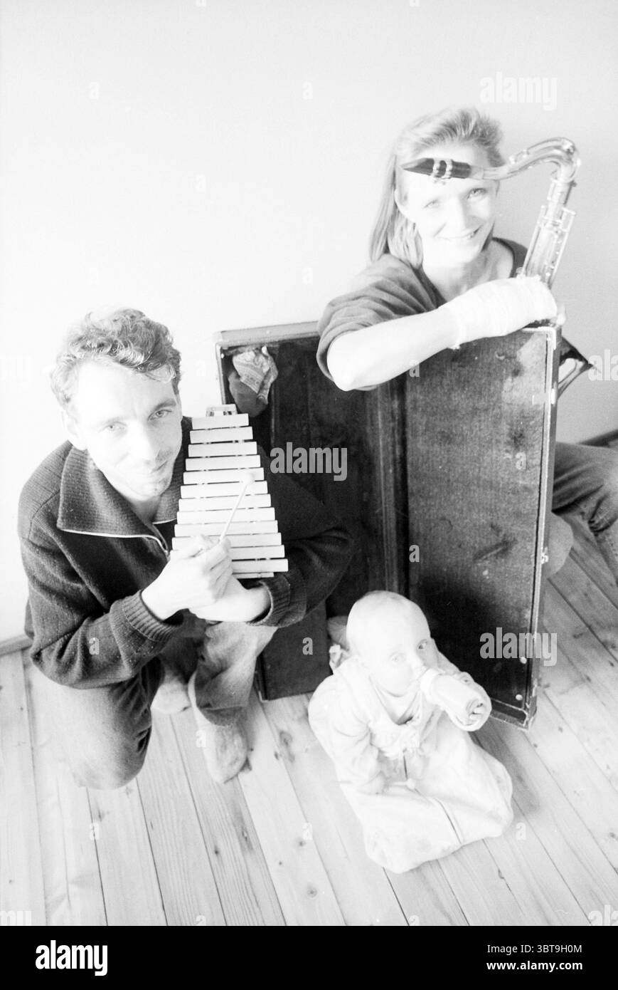 Two people from the Haarlem Street Orchestra Haarlem The Netherlands, Whizgle News, Dutch Desk, The Netherlands, 1950 - 2000 on 24-04-1992. These topics appear in the image. In a dimly lit room, two adults and a child create a captivating scene. The adults sit closely, each exuding a sense of personality and artistry. One, with tousled hair, holds a wooden xylophone, carefully structured with its bars arranged in a cascading pattern. The other, a woman with striking hair, is poised in a unique pose, holding a saxophone while resting against a large, vintage case that adds a sense of nostalgia Stock Photo