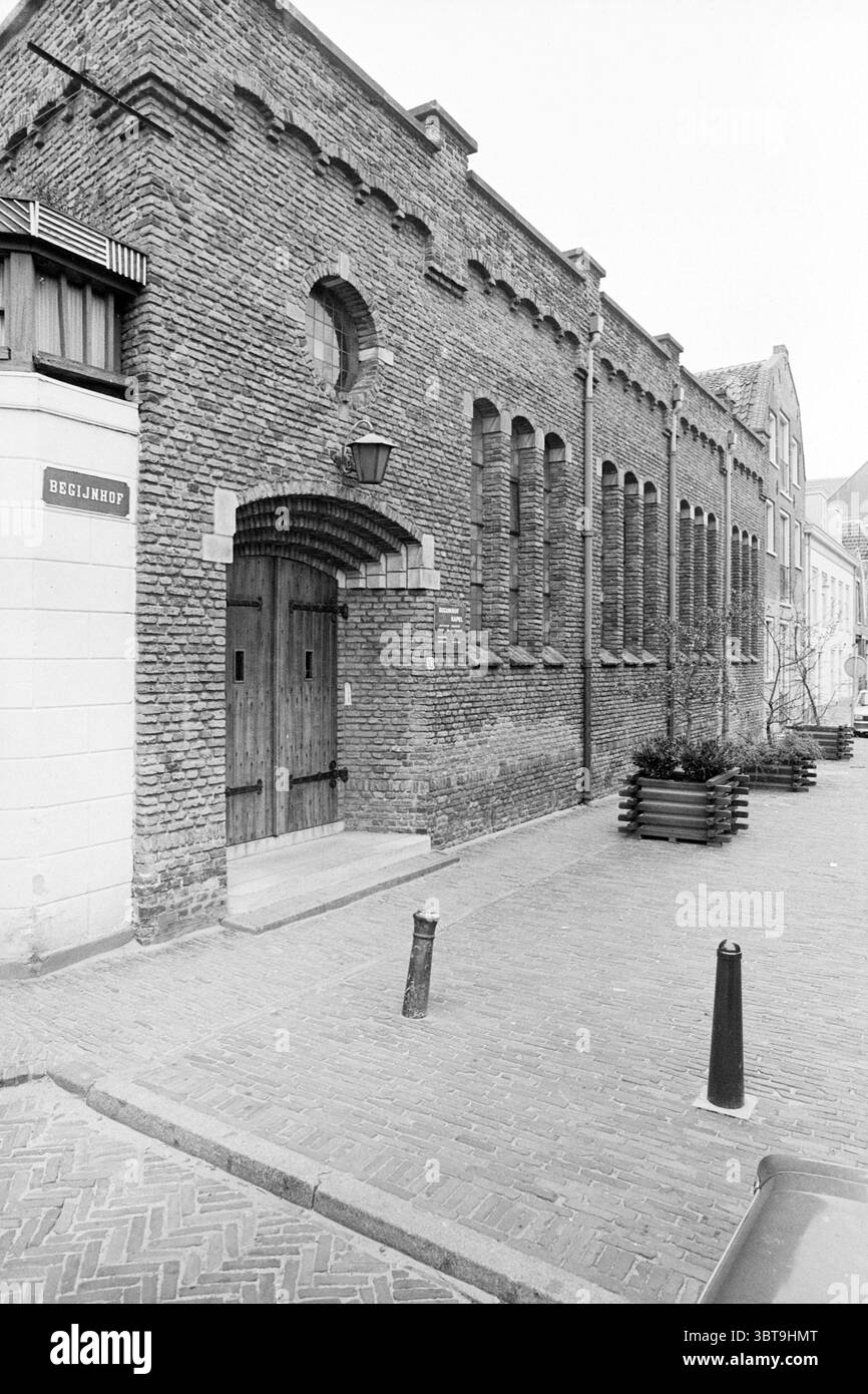 Exterior Baptist Chapel Beguinage Churches Haarlem Begijnhof The Netherlands, Whizgle News, Dutch Desk, The Netherlands, 1950 - 2000 on 04-11-1980. The image contains these topics. The scene depicts a street lined with robust brick buildings, characterized by a textured facade that suggests a sense of enduring strength. The dominant colors are muted tones of gray and brown, contributing to an overall somber yet timeless aesthetic. Among the buildings, one features an imposing wooden door framed by intricate brickwork, with pointed arches above that enhance its historic feel. On the left, a pol Stock Photo