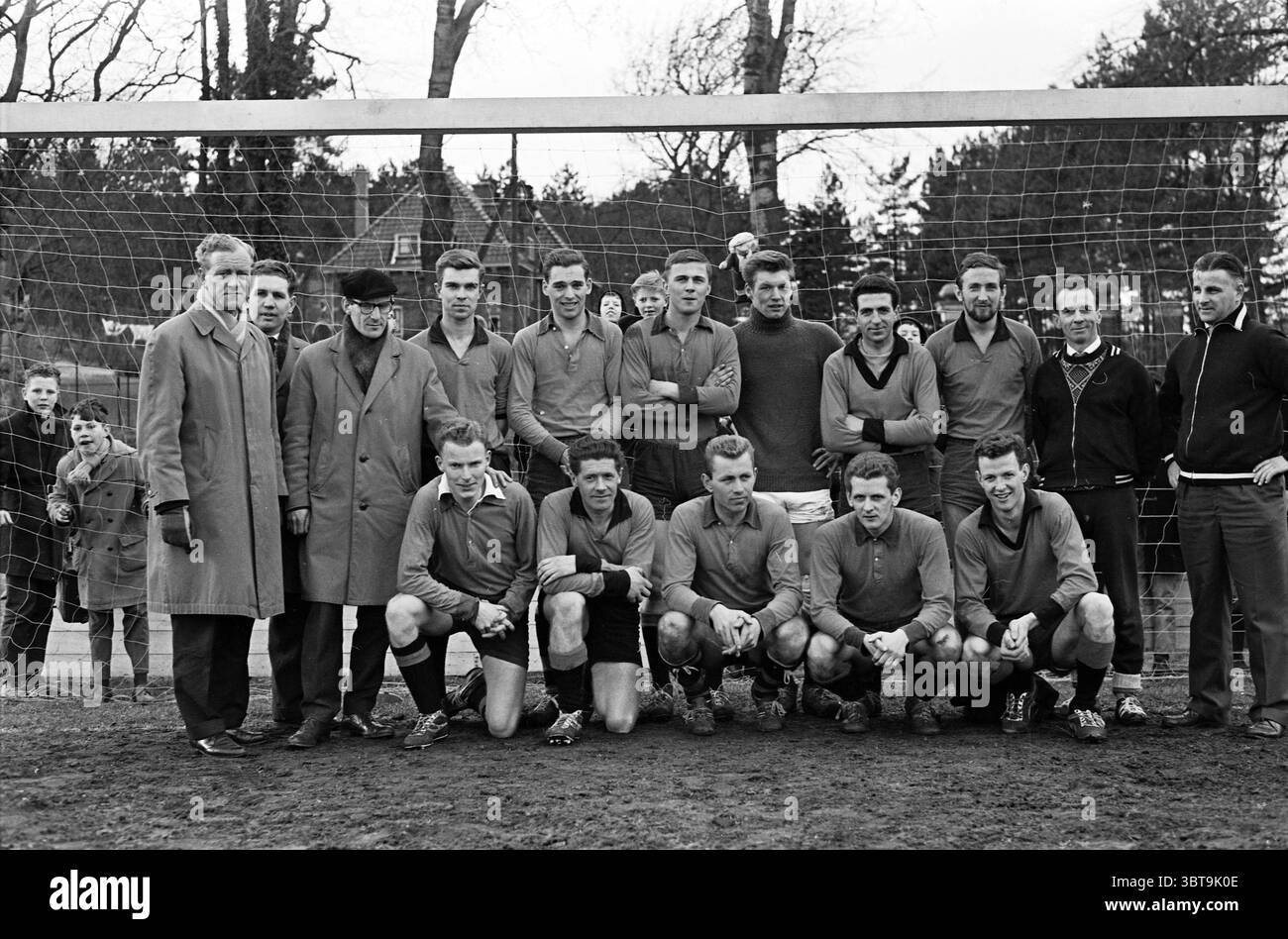 Kennemerland Teams Football, Whizgle News, Dutch Desk, The Netherlands, 1950 - 2000 on 24-03-1962. The image includes these topics. The scene captures a vintage team photo of a football (soccer) squad, positioned in front of a goalpost. The players are arranged in two rows, displaying a mix of confident smiles and focused expressions. In the front row, players are kneeling, wearing matching t-shirts in a muted tone, likely a shade of blue or gray, with shorts and football socks, exuding a sense of camaraderie and unity. The back row features standing players, dressed similarly, with varying de Stock Photo