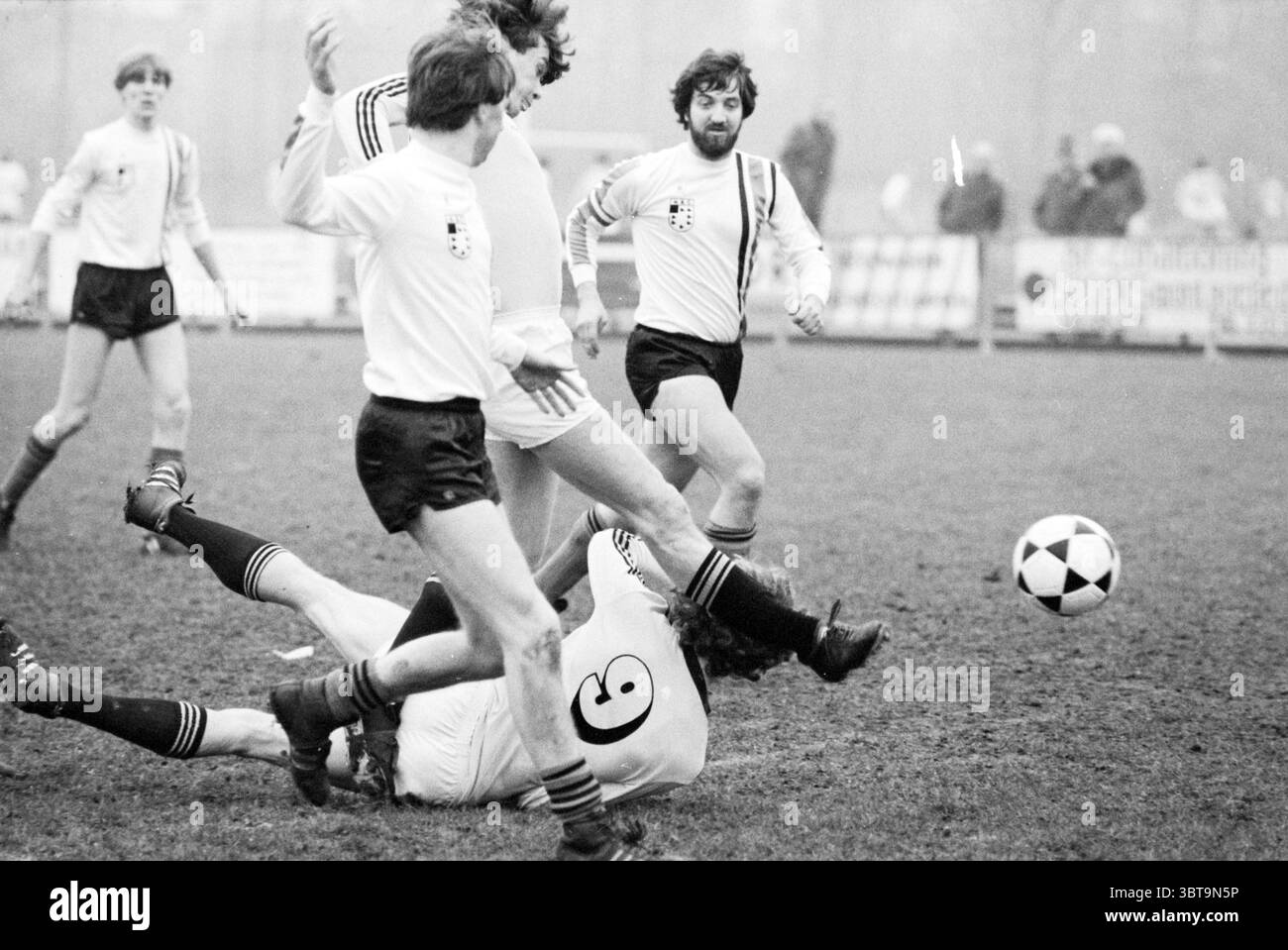 NAC football match, Whizgle News, Dutch Desk, The Netherlands, 1950 - 2000 on 01-1981. These are the topics in the image. The scene captures a dynamic moment during a soccer match, populated by players in motion. Several individuals wearing classic white jerseys with black shorts are engaged in an intense play. Their uniforms have a vintage appearance, characterized by a simple design that hints at a bygone era of the sport. The grassy field is slightly muddy, suggesting recent rainfall or heavy use, contributing to the overall earthy tones of the scene. The players, with a mix of determinatio Stock Photo