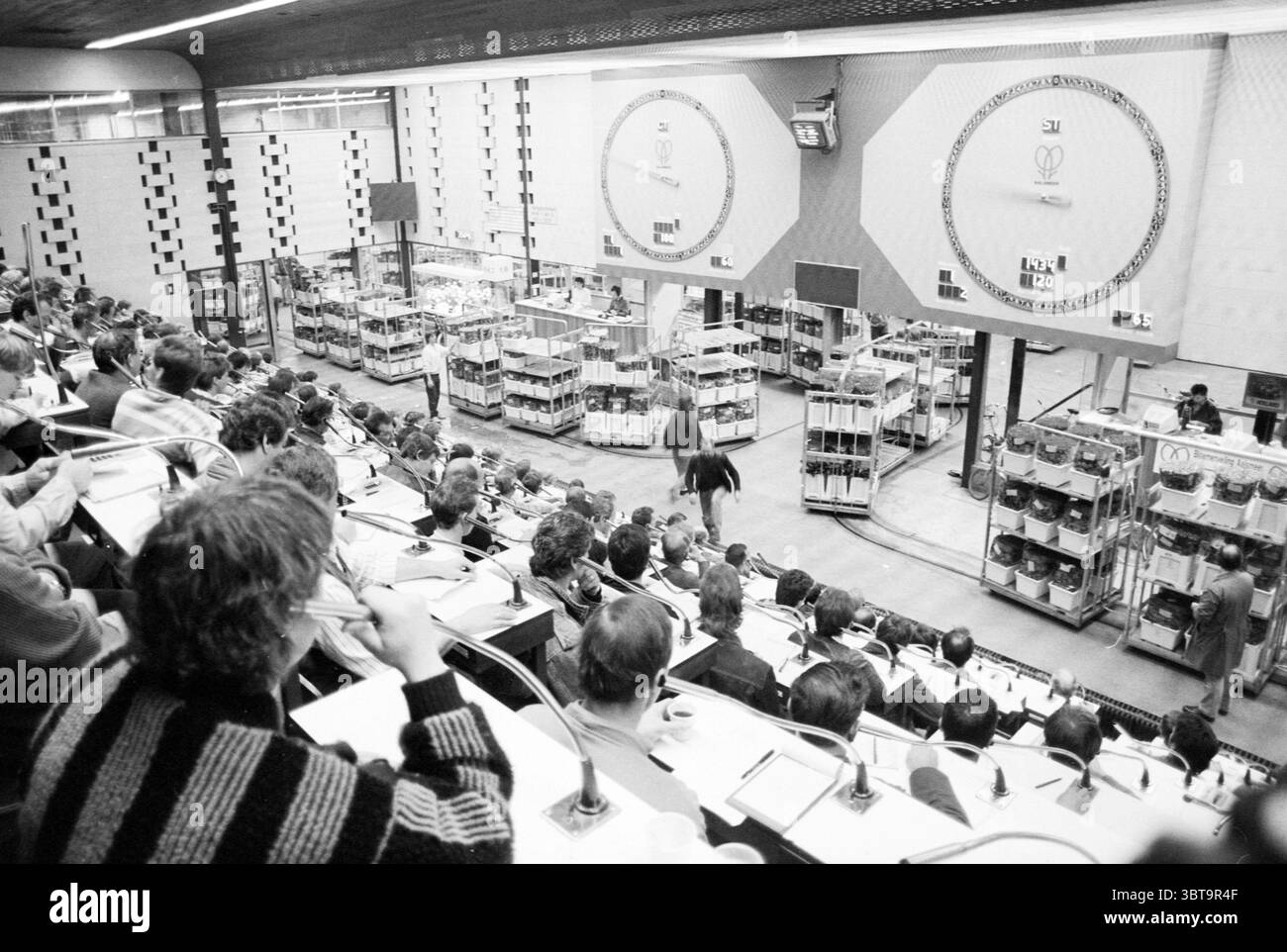 Flower Auction Aalsmeer Auction Aalsmeer, Whizgle News, Dutch Desk, The Netherlands, 1950 - 2000 on 30-10-1987. The image shows these topics. The scene captures a bustling trading floor filled with individuals engaged in intense discussion and observation. Rows of people are seated in a tiered configuration, focusing on the activities in the center of the room. Many are dressed in professional attire, suggesting a formal atmosphere conducive to business dealings. The setting has a modern feel, accentuated by clean lines and a somewhat industrial aesthetic, with metal and glass elements promine Stock Photo