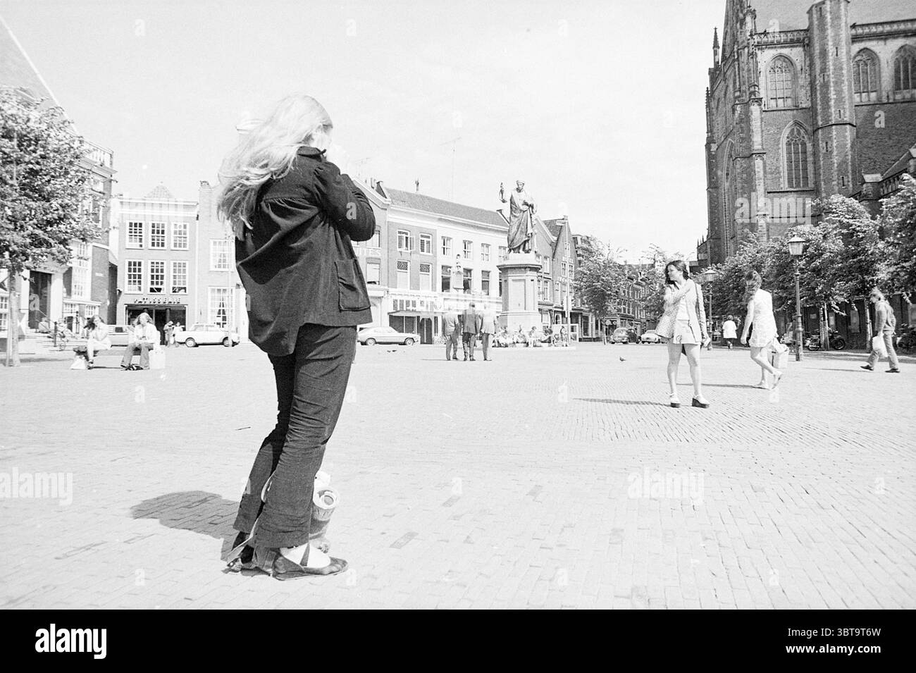 Report Loutje Grote Markt Report, Whizgle News, Dutch Desk, The Netherlands, 1950 - 2000 on 09-08-1972. These are the elements in the image. In a lively urban square bathed in bright daylight, a woman with long, flowing blonde hair stands prominently in the foreground. She is dressed in a black ensemble that contrasts sharply with the lighter tones of her surroundings. Her attention is directed towards something in front of her, perhaps capturing the scene through a camera or observing the activity around her. The square is paved with cobblestones, and its expansive space allows for a variety Stock Photo