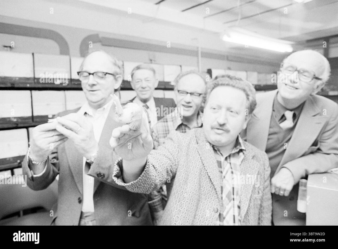 Bird exchange fair IJmuiden Birds, Whizgle News, Dutch Desk, The Netherlands, 1950 - 2000 on 17-11-1978. The image shows these topics. The scene captures a group of five individuals gathered closely together in an indoor setting, likely a laboratory or a workshop. The atmosphere appears lively and engaging, with the group focused on a specific object being held up by one of the men at the forefront. The man in the center, holding up his hand, shows a distinct gesture, perhaps pointing or displaying something to the others. His attire features a patterned blazer, giving a sense of personality a Stock Photo