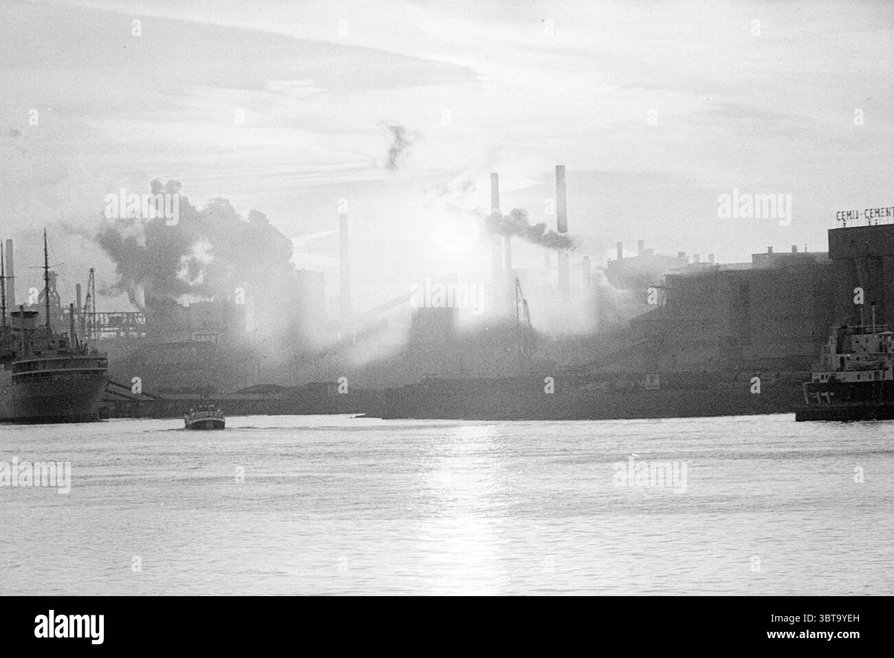 Atmosphere photo Sunset Blast Furnaces IJmuiden Atmosphere, Whizgle News, Dutch Desk, The Netherlands, 1950 - 2000 on 18-06-1973. These are the elements in the image. The scene depicts a bustling industrial landscape at dusk. In the foreground, a serene body of water reflects the gray hues of the sky, which is filled with soft, diffused light. Several boats are anchored, their silhouettes stark against the glimmering surface. On the horizon, a sprawling industrial complex looms, marked by towering smokestacks releasing plumes of dark, swirling smoke that merge with the low-hanging clouds. The Stock Photo