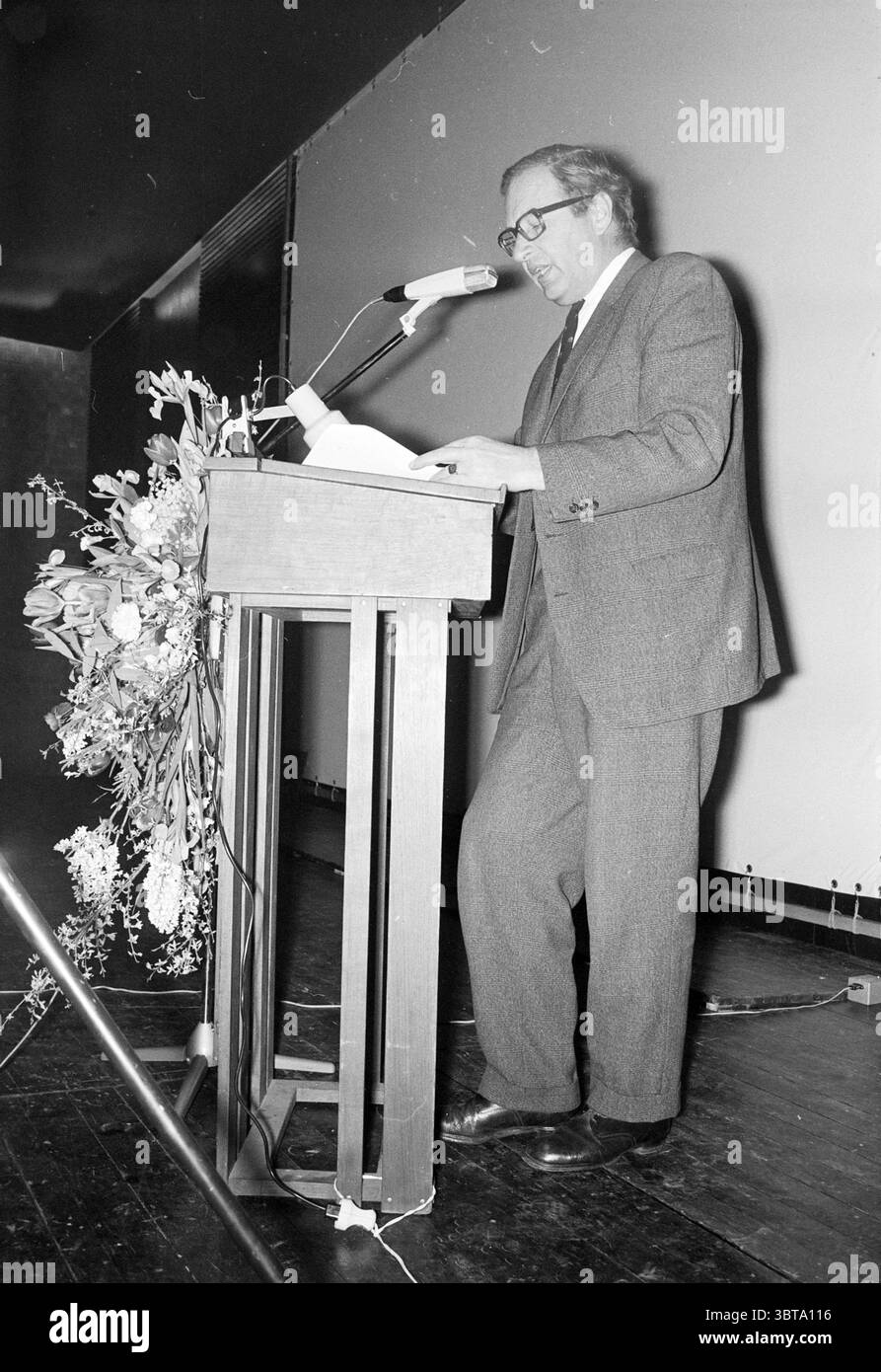 Speech Mr. de Jonge safety roll call Hoogovens. Hoogovens, Whizgle News, Dutch Desk, The Netherlands, 1950 - 2000 on 05-02-1975. These are the elements in the image. The scene captures a formal setting with an individual standing behind a wooden lectern, which is designed for speaking engagements. The lectern is elegantly simple, featuring a polished surface and a small floral arrangement that adds a touch of decorum. The person is dressed in a tailored suit, characterized by subtle patterns, and is wearing glasses that reflect an air of intellect. They are engaged in delivering a speech, with Stock Photo