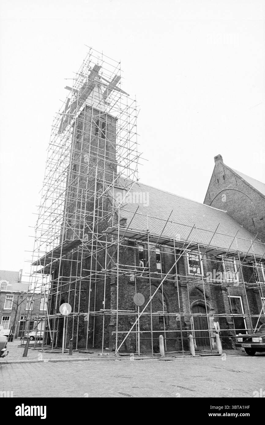 Exterior Walloon church in scaffolding Haarlem Begijnhof The Netherlands, Whizgle News, Dutch Desk, The Netherlands, 1950 - 2000 on 12-03-1981. These topics are shown in the image. The scene features a tall church building, primarily constructed of dark, weathered brick, which emphasizes its historical significance. Scaffolding envelops the structure, creating a stark contrast with the timeworn materials of the building. The scaffolding is a mix of metal and wood, showcasing interwoven beams that support the renovation process. The church has a distinctive tower that rises prominently, reachin Stock Photo