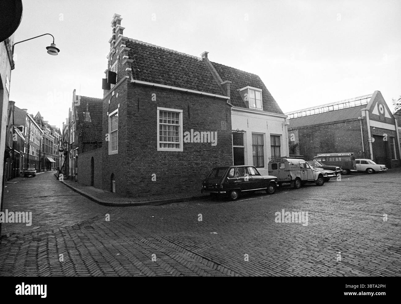 Begijnhof building Exterior Haarlem Begijnhof The Netherlands, Whizgle News, Dutch Desk, The Netherlands, 1950 - 2000 on 15-09-1972. The image shows these topics. The scene showcases a narrow, cobblestone street corner, creating a quaint ambiance. On one side, a distinctive brick building stands prominently, characterized by its gabled roof and a large window that allows glimpses of the interior. Its earthy red tones contrast with the muted gray sky overhead, which suggests an overcast day. Flanking the building, several vehicles are parked along the street—each a different model and color, co Stock Photo