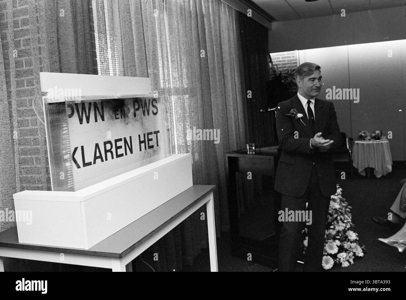 Opening of the provincial water company laboratory Opening openings putting into use, Whizgle News, Dutch Desk, The Netherlands, 1950 - 2000 on 28-05-1980. The image includes these topics. A well-lit indoor setting features a man standing confidently, wearing a dark suit accented with a boutonniere. He appears engaged, possibly addressing an audience or making a presentation. The backdrop is adorned with draped curtains in soft, muted tones, creating a formal atmosphere. In front of him, a transparent display case rests on a simple white pedestal, holding a notable object that draws attention, Stock Photo