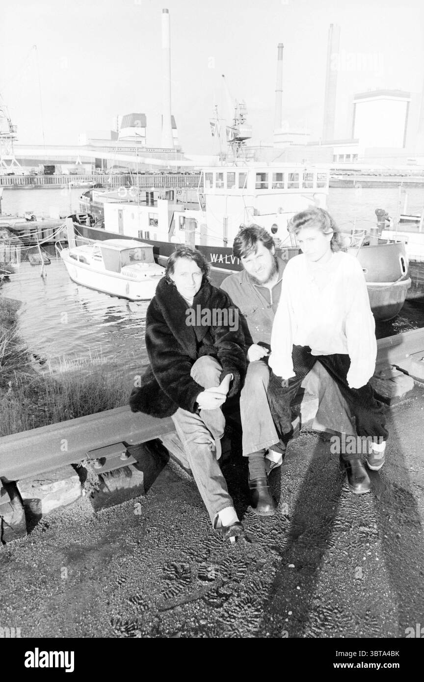 Family Hendrikse ship the Walvis Ships, Whizgle News, Dutch Desk, The Netherlands, 1950 - 2000 on 28-01-1986. These are the elements in the image. In a stark, monochromatic setting, three individuals are positioned prominently in the foreground, sitting casually along a sturdy, industrial barrier that runs alongside a body of water. The scene has a gritty, urban feel, characterized by the presence of moored boats in the background, which reflect a mix of shapes and sizes. The people appear relaxed, perhaps engaging in conversation or contemplating the view. One person is draped in a dark, plus Stock Photo