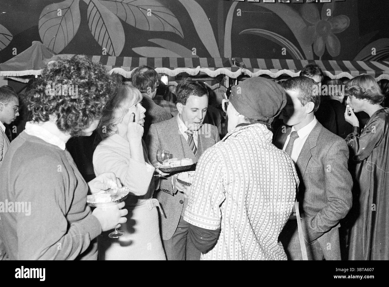 Party 25th anniversary Fa. Abraham Kef cheese trader fromagerie Amsterdam The Netherlands, Whizgle News, Dutch Desk, The Netherlands, 1950 - 2000 on 31-10-1980. The image contains these topics. In a lively social gathering, the scene presents a mix of individuals engaged in conversation and interaction. People are dressed in a combination of formal and semi-formal attire, indicating a festive atmosphere. The central figure holds a tray, possibly balanced with drinks or appetizers, while engaging with two companions. One of them, a woman, has an elegant hairstyle and is seen gesturing, perhaps Stock Photo