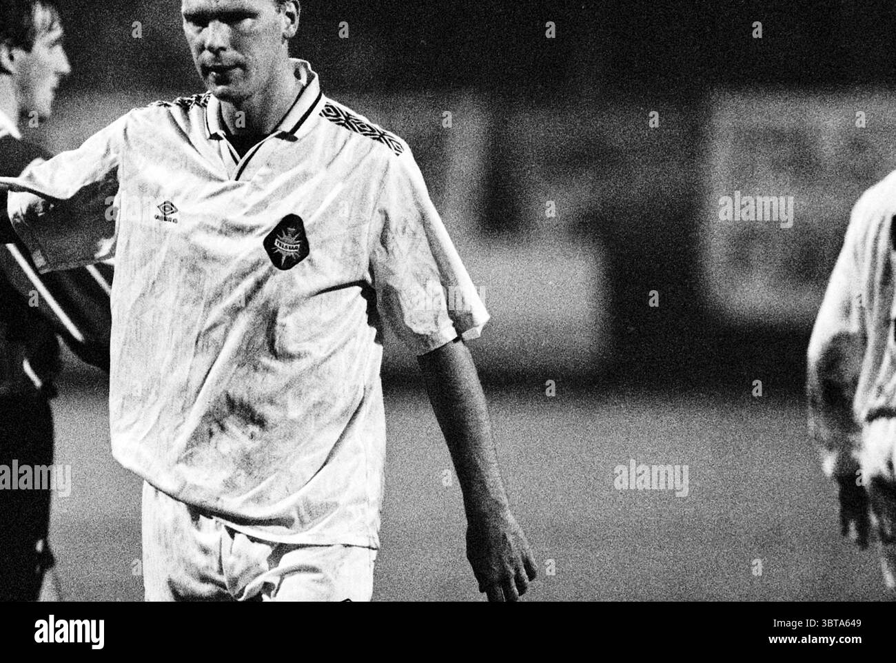 Football Telstar - Emmen, Whizgle News, Dutch Desk, The Netherlands, 1950 - 2000 on 27-10-1995. These are the elements in the image. The scene captures a moment in a sporting event, likely during a soccer match. The player in the foreground wears a loose-fitting, white jersey adorned with a simple emblem on the chest, characterized by an understated, vintage design. The jersey appears slightly wrinkled, possibly from the exertion of the game. His shorts match the jersey in color, and he looks focused yet fatigued, suggesting a recent contest on the field. The player's short hair and determined Stock Photo