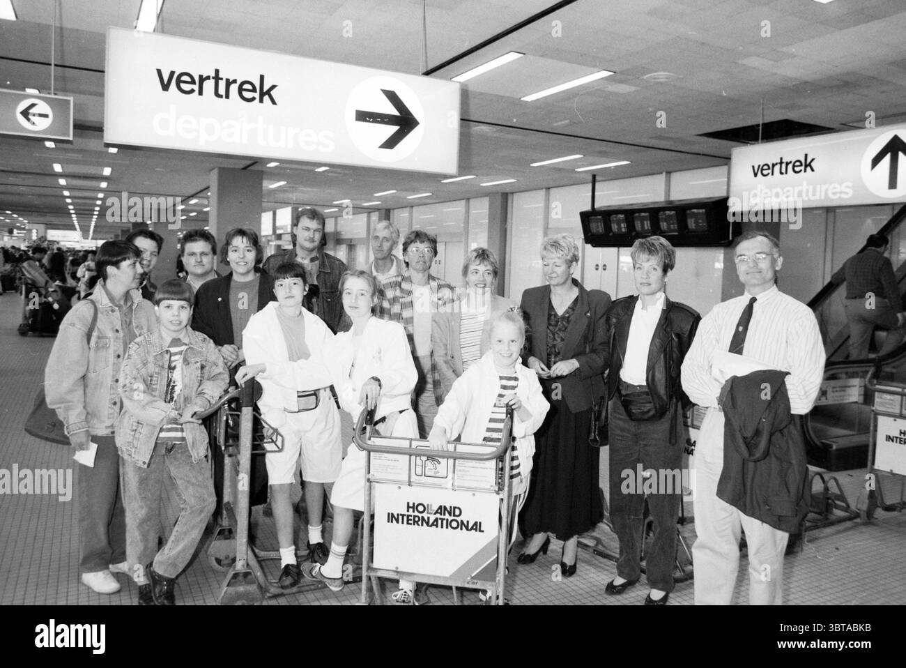 Prize winners from IJmuiden Schiphol Schiphol, Whizgle News, Dutch Desk, The Netherlands, 1950 - 2000 on 12-04-1991. These are the topics in the image. In a bustling airport terminal, a diverse group of people gathers in front of a large sign indicating the direction of departures. The scene is characterized by a mix of emotions, from excitement to a hint of apprehension, as faces express a range of sentiments. The individuals are casually dressed, blending various styles, indicating that they come from different backgrounds. Some are in comfortable travel attire, like T-shirts and jeans, whil Stock Photo
