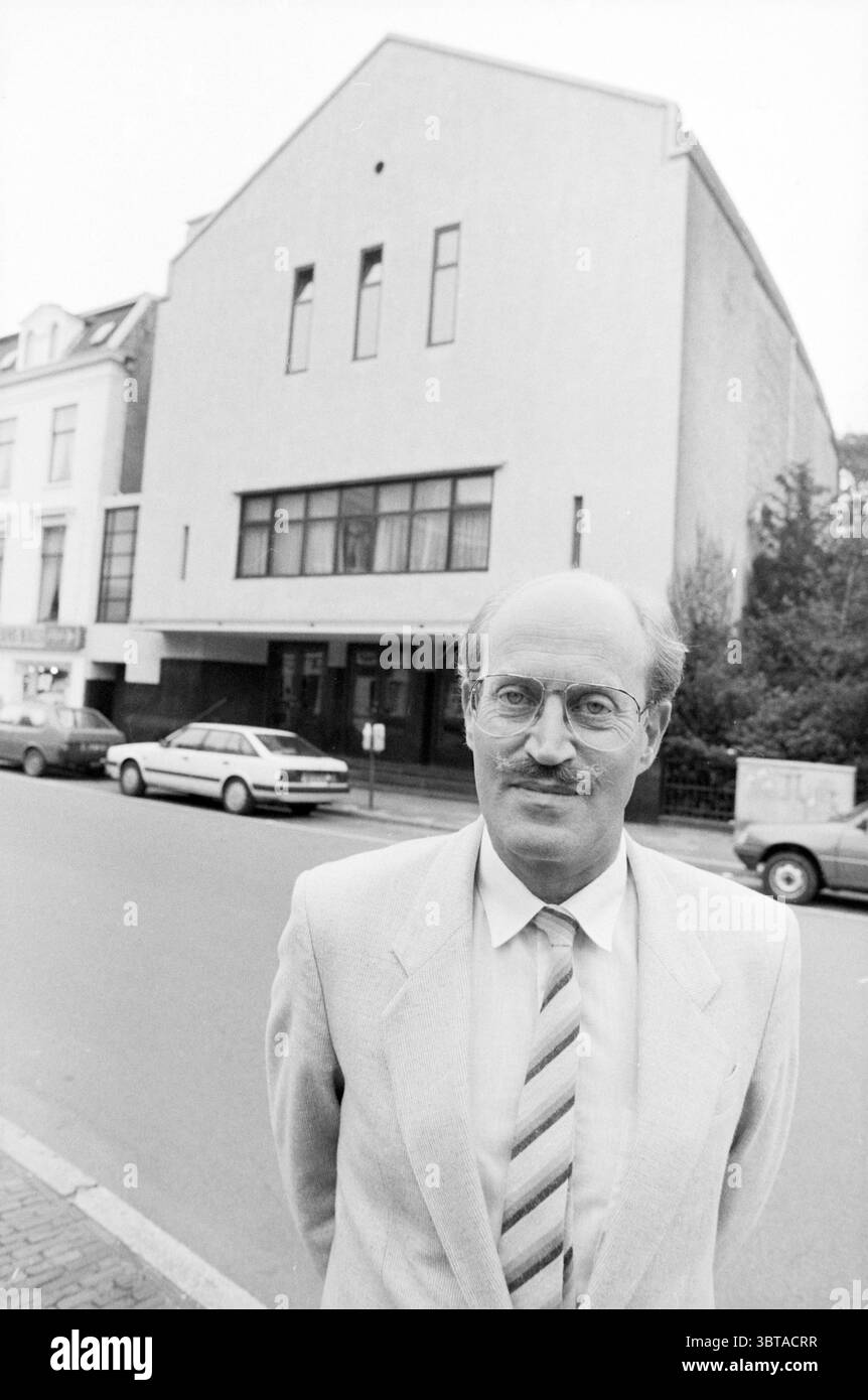 Mr. van de Burg former Frans Hals cinema Persons, Whizgle News, Dutch Desk, The Netherlands, 1950 - 2000 on 02-09-1987. These topics are shown in the image. In the scene, a middle-aged man stands prominently in the foreground, exuding a calm demeanor. He wears a light-colored suit with a striped tie that introduces subtle hints of color—maybe shades of blue and beige. His hair is light and neatly combed, and he has a thin mustache that complements his thoughtful expression. Behind him, a large, modern building with a distinct architectural style imposes itself. Its exterior showcases a mostly Stock Photo
